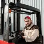 Portrait of a forklift operator inside a warehouse, wearing a uniform and cap.