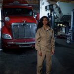 A mechanic wearing a jumpsuit stands confidently in a garage with a red truck.